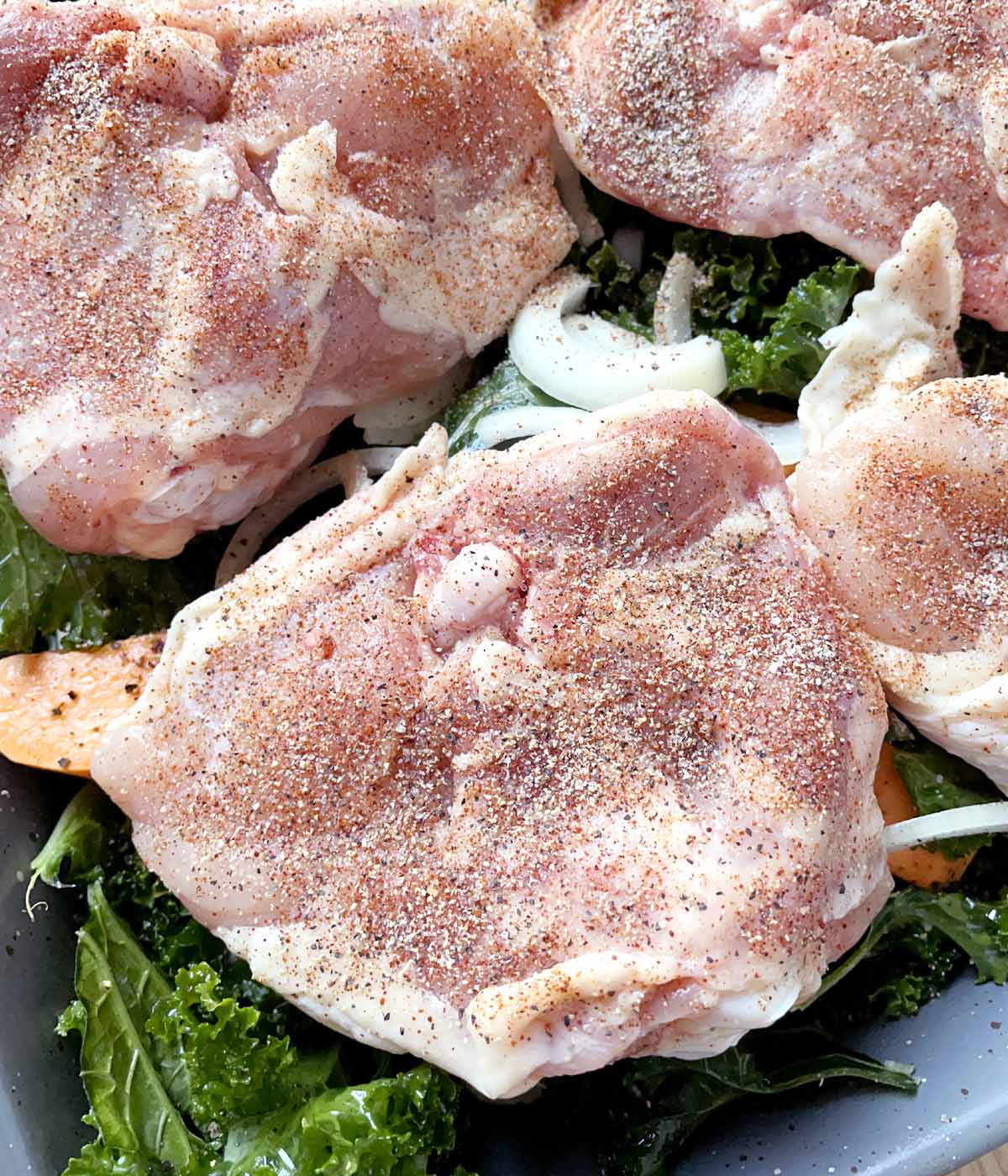 Raw chicken pieces with dry seasonings on them, resting on a bed of uncooked vegetables in a roasting pan.