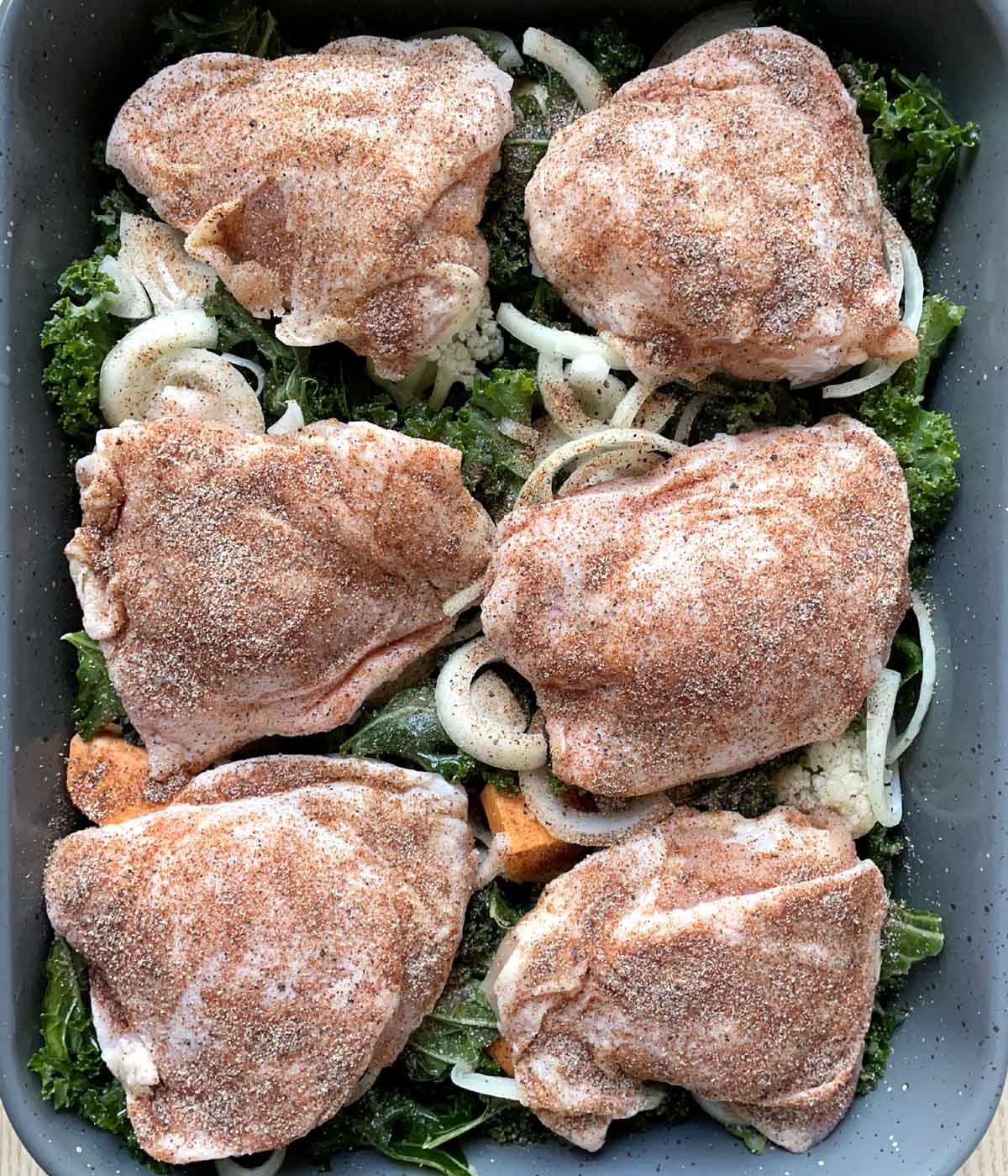 Six raw chicken pieces on a bed of uncooked vegetables in a rectangular roasting pan.