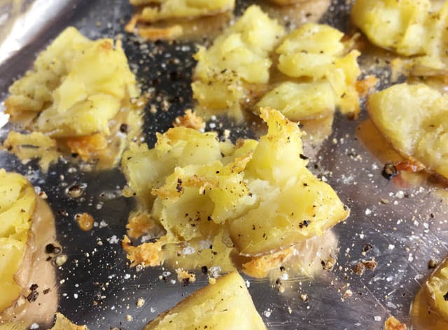 Crispy roasted smashed potatoes surrounded by salt and pepper on a foil-lined baking sheet