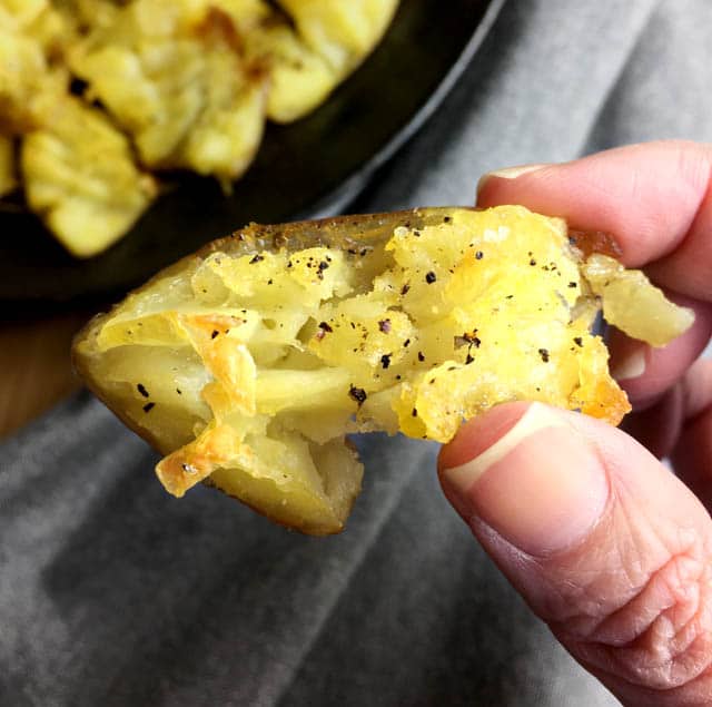 Close-up of fingers holding crispy roasted smashed potatoes with crisp edges and black pepper specks