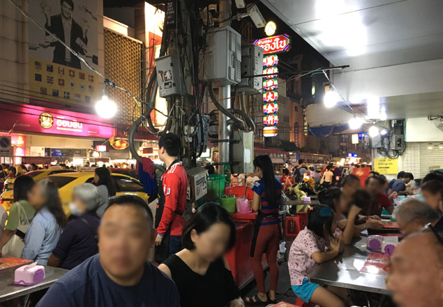 A crowd of people sitting next to a busy street in Chinatown, Bangkok