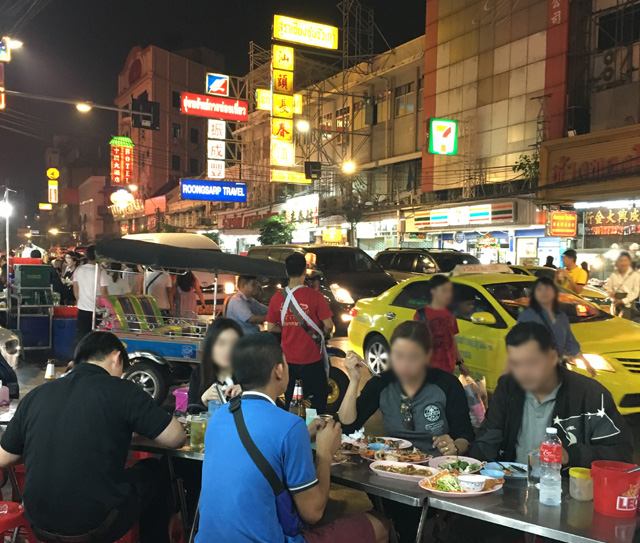 People dining at tables next to a busy road full of cars in Chinatown, Bangkok