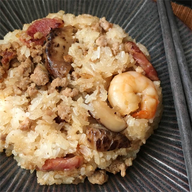 A dark round plate containing rice mixed with mushrooms, shrimp, and red Chinese sausage