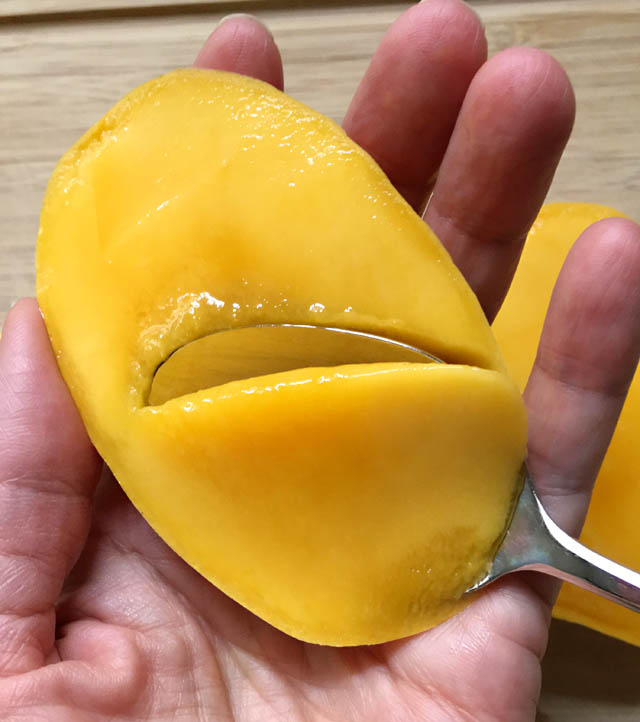 Close-up of a hand holding a section of mango and a spoon scooping out the mango flesh