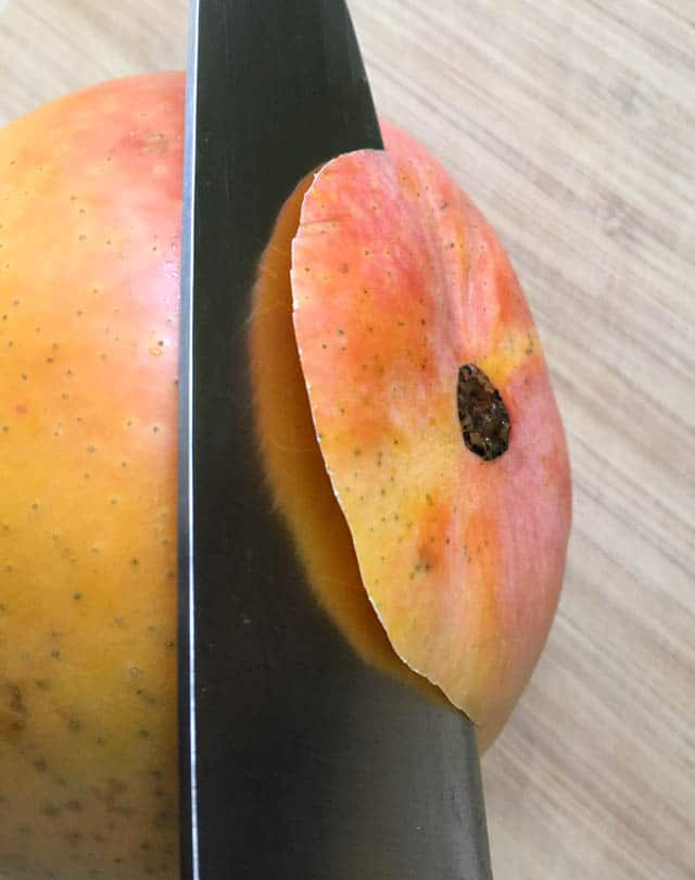 Close-up of a knife slicing through the bottom of a yellow orange mango on a wooden cutting board