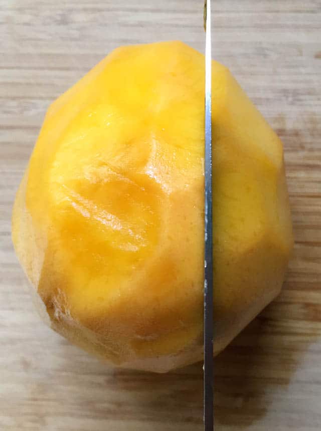 A knife slicing into a large yellow orange mango on a wooden cutting board