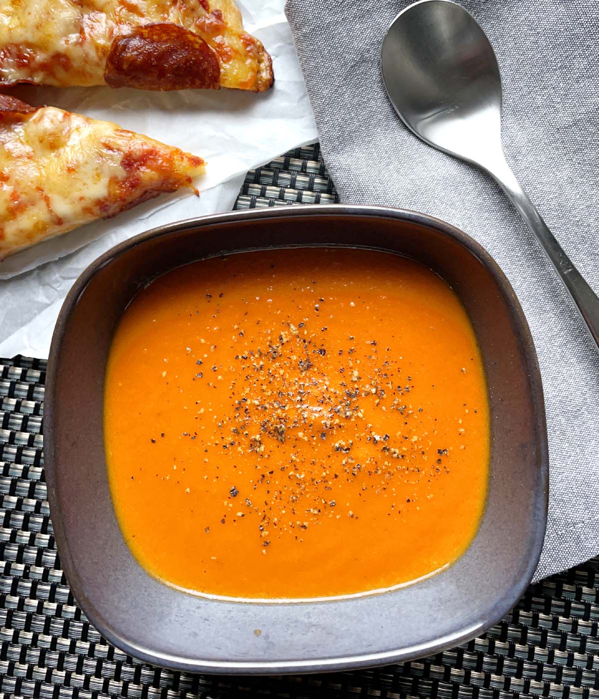 A squarish grey bowl containing roasted tomato soup, next to a spoon and a couple slices of pizza.