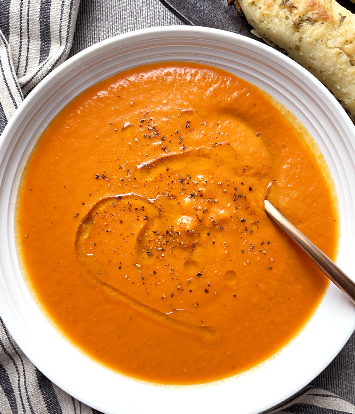 A round white bowl containing orange roasted tomato soup topped with a drizzle of oil and cracked black pepper.