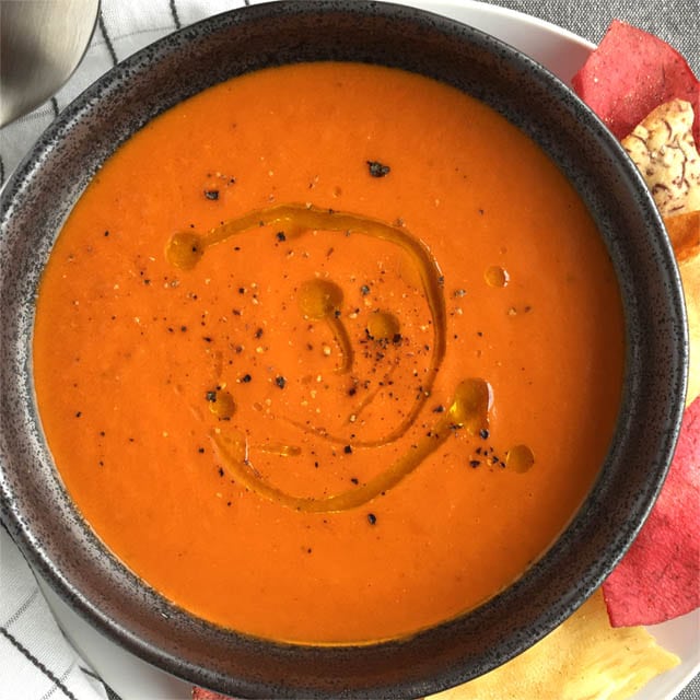 A dark round bowl containing orange roasted tomato soup with olive oil drizzle and cracked black pepper on top