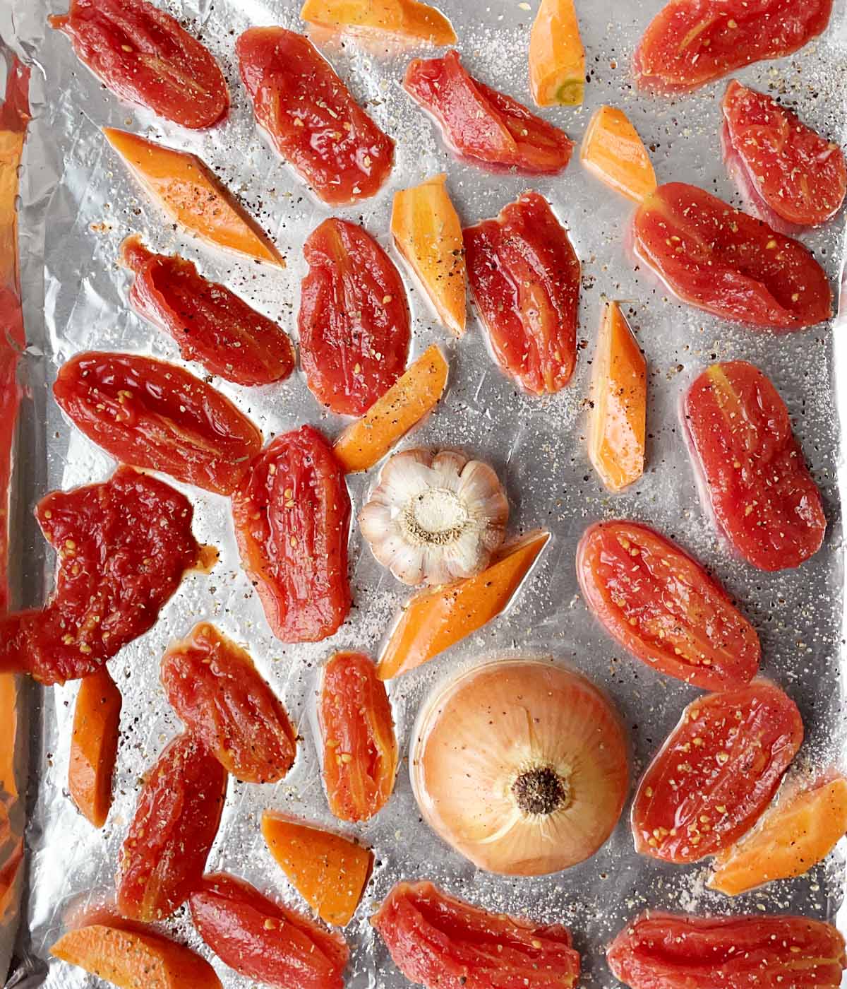 Sliced red tomatoes, carrot chunks, a bulb of garlic, and half an onion on a foil-lined baking sheet.