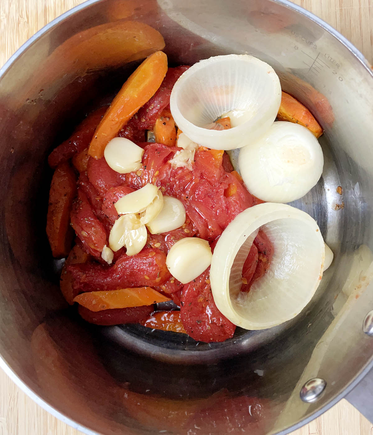 A metal pot containing red tomatoes, carrots, onions, and garlic.