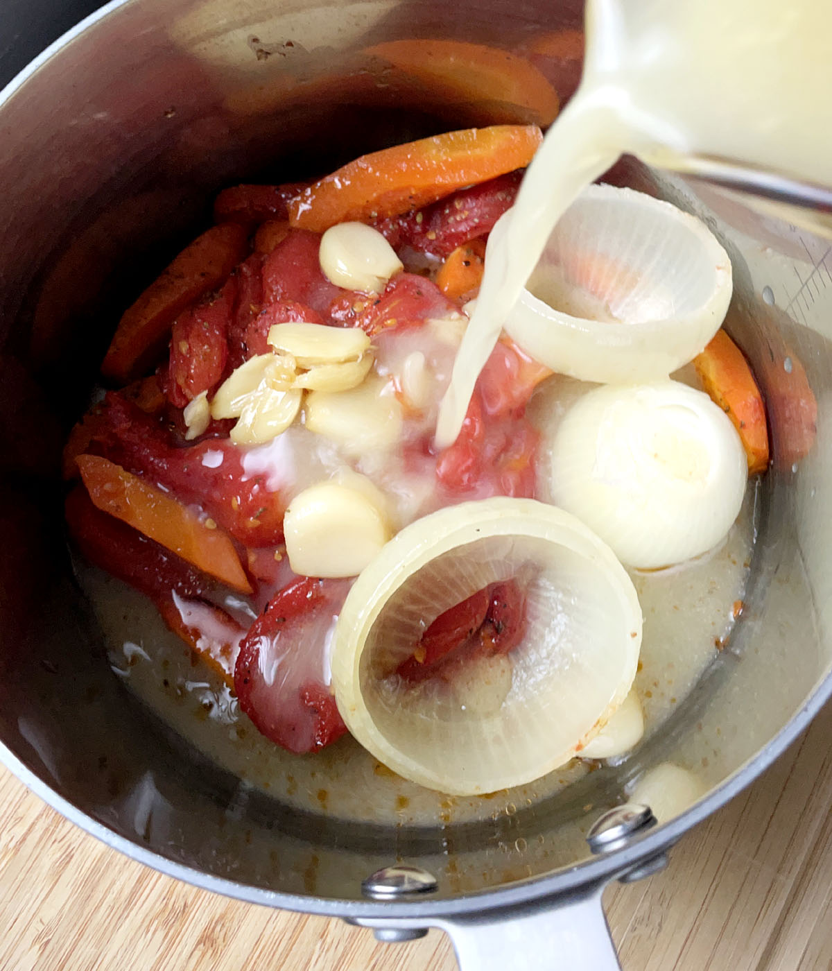 Broth being poured into a pot containing tomatoes, onions, garlic, an carrots.