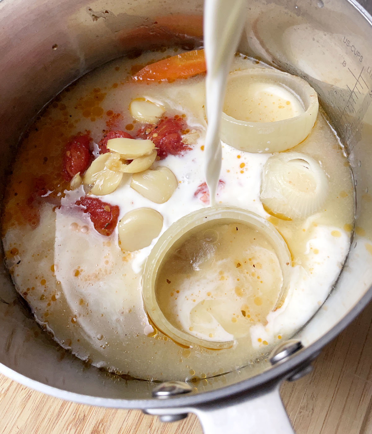 White milk being poured into a pot containing onions, garlic, and tomatoes.
