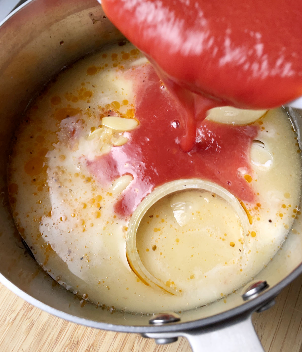 Red tomato sauce being poured into a pot containing onions and a milky liquid.