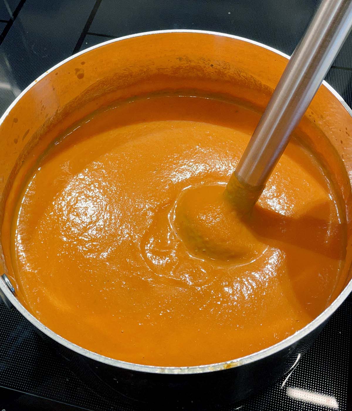 A metal immersion blender in a pot filled with orange roasted tomato soup.