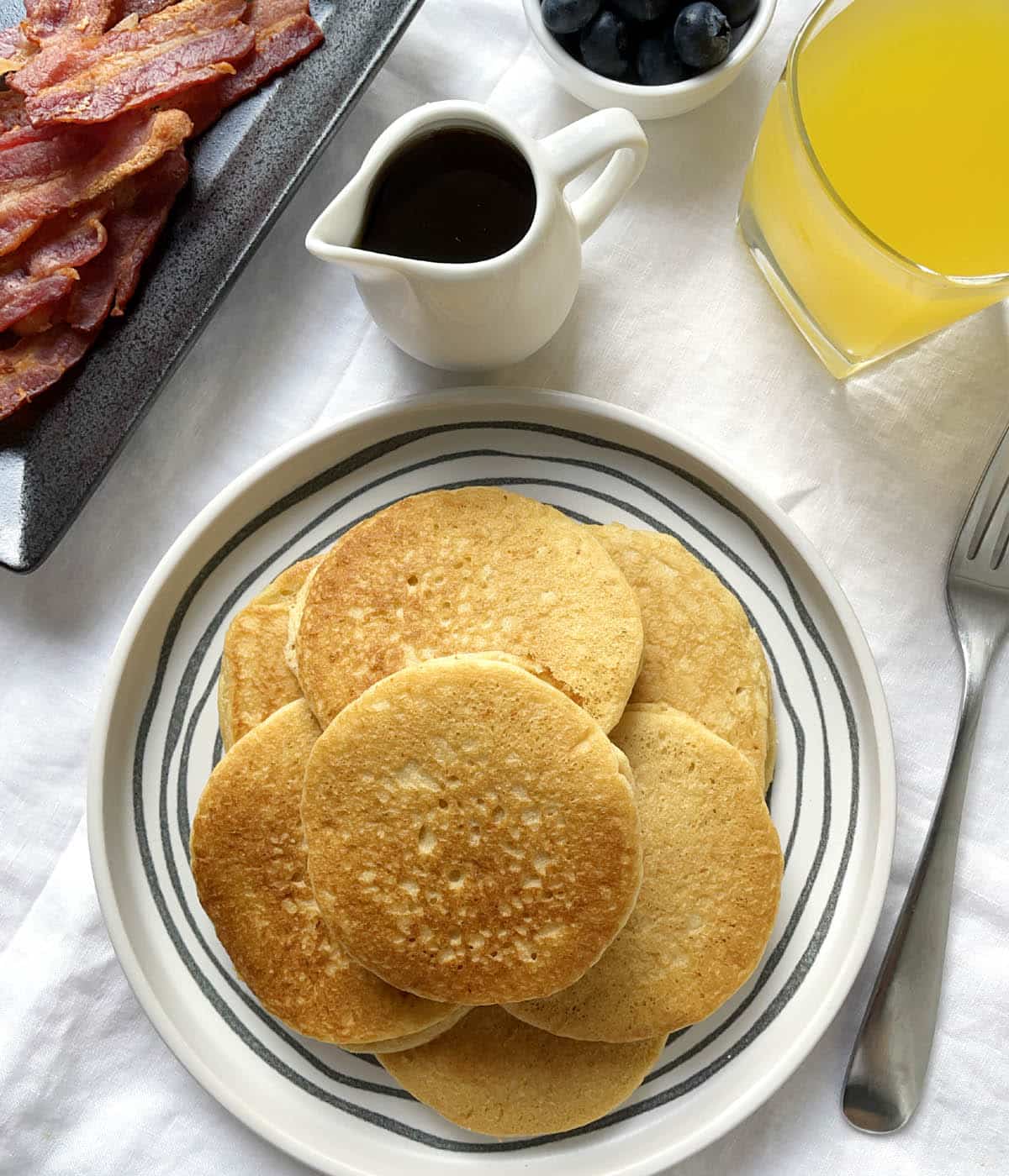 A white round plate containing almond pancakes, next to a plate of bacon, a pitcher of syrup, a glass of orange juice, and a fork.
