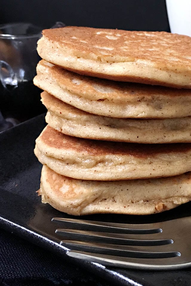 Close-up of a fork in front of a stack of 5 brown almond pancakes on a black plate