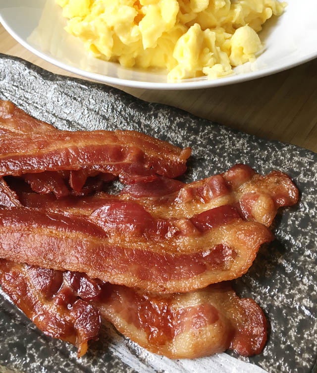 A grey plate containing slices of cooked bacon for a crowd next to a white bowl containing yellow scrambled eggs