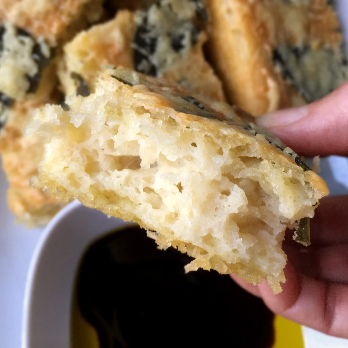 Close-up of the inside of a piece of parmesan sage focaccia bread being help by a hand over a bowl of oil and dark brown vinegar.