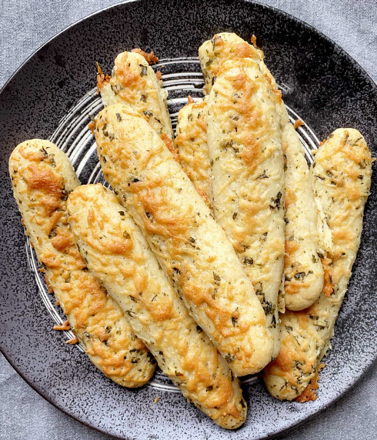 A dark round dish containing eight baked parmesan breadsticks.