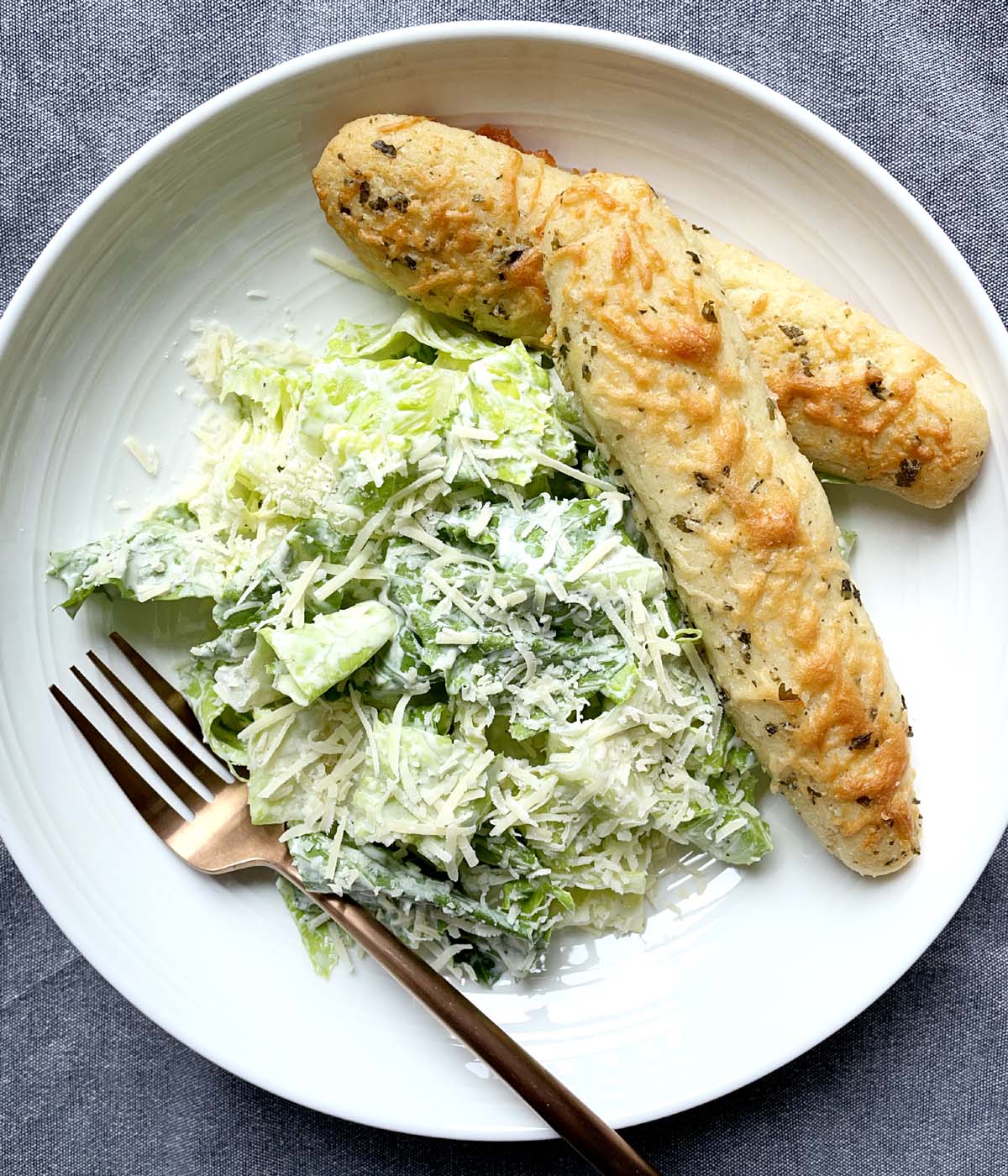 A white round dish containing a brown metal fork, Caesar salad, and two breadsticks.