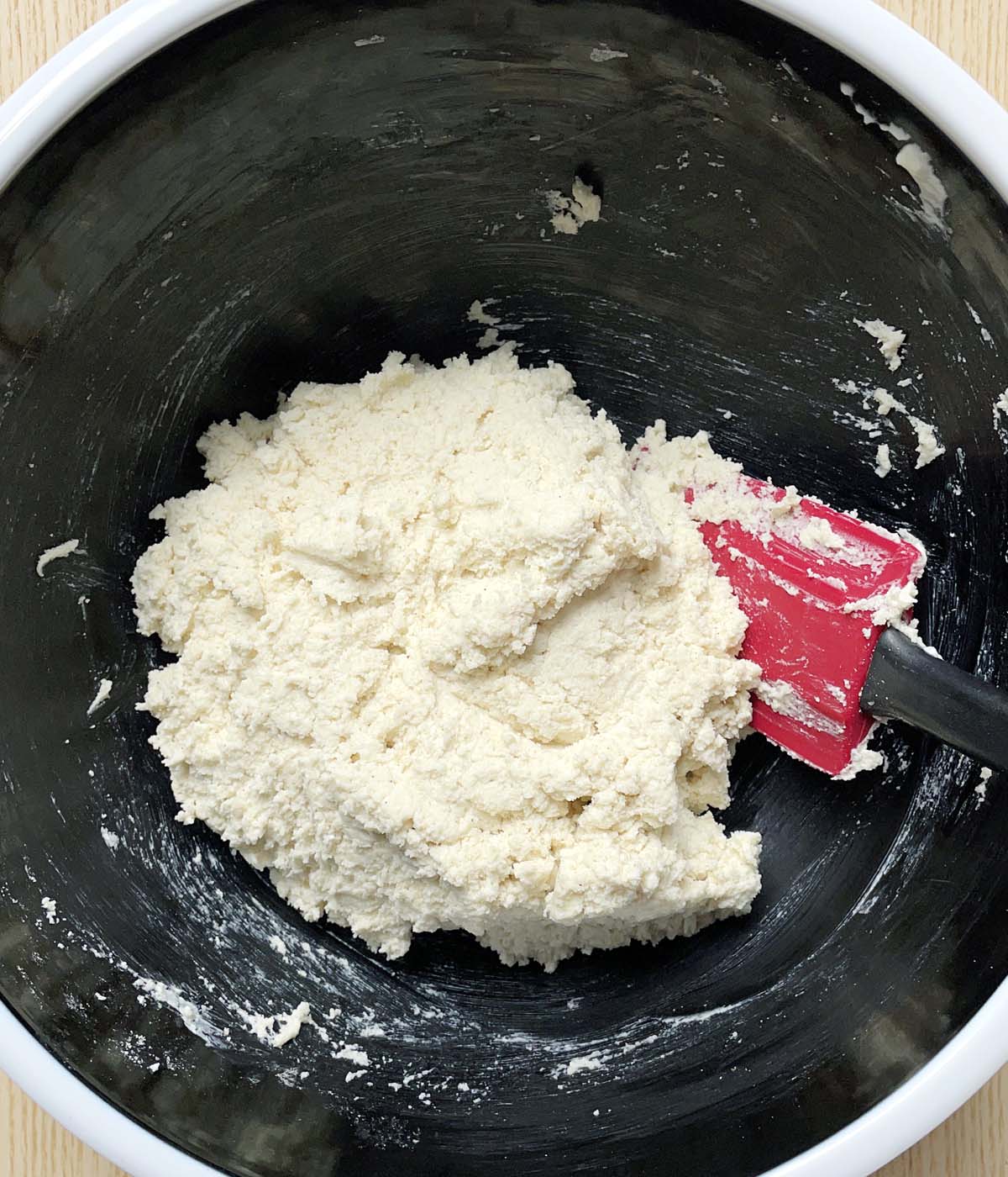 A ball of shaggy light colored dough and a red rubber spatula in a black bowl.