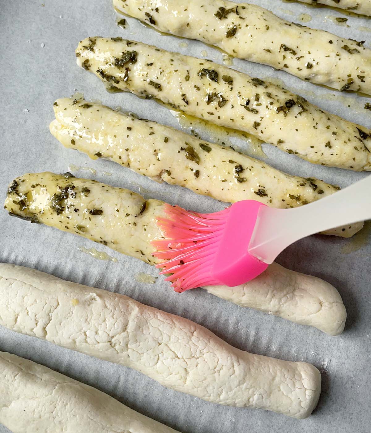 Melted butter with parsley bits being brushed on breadstick dough with a pink brush.
