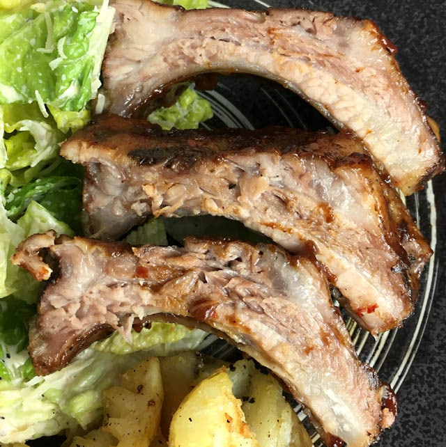 Three pork ribs on a dark plate containing green salad and yellow potatoes