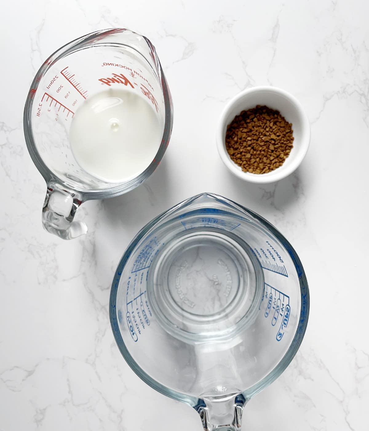 A measuring cup containing water, a measuring cup containing white milk, and white round dish containing brown coffee granules.