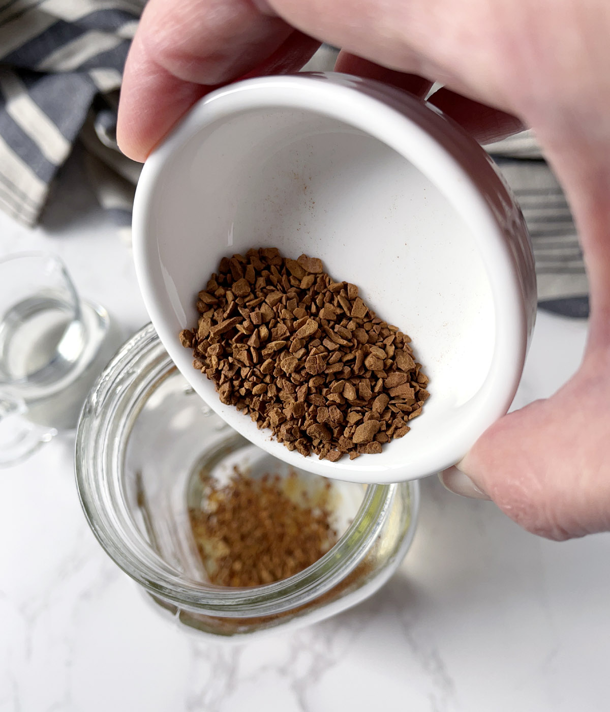 Close-up of a hand holding a white dish pouring brown coffee granules into a glass bottle.
