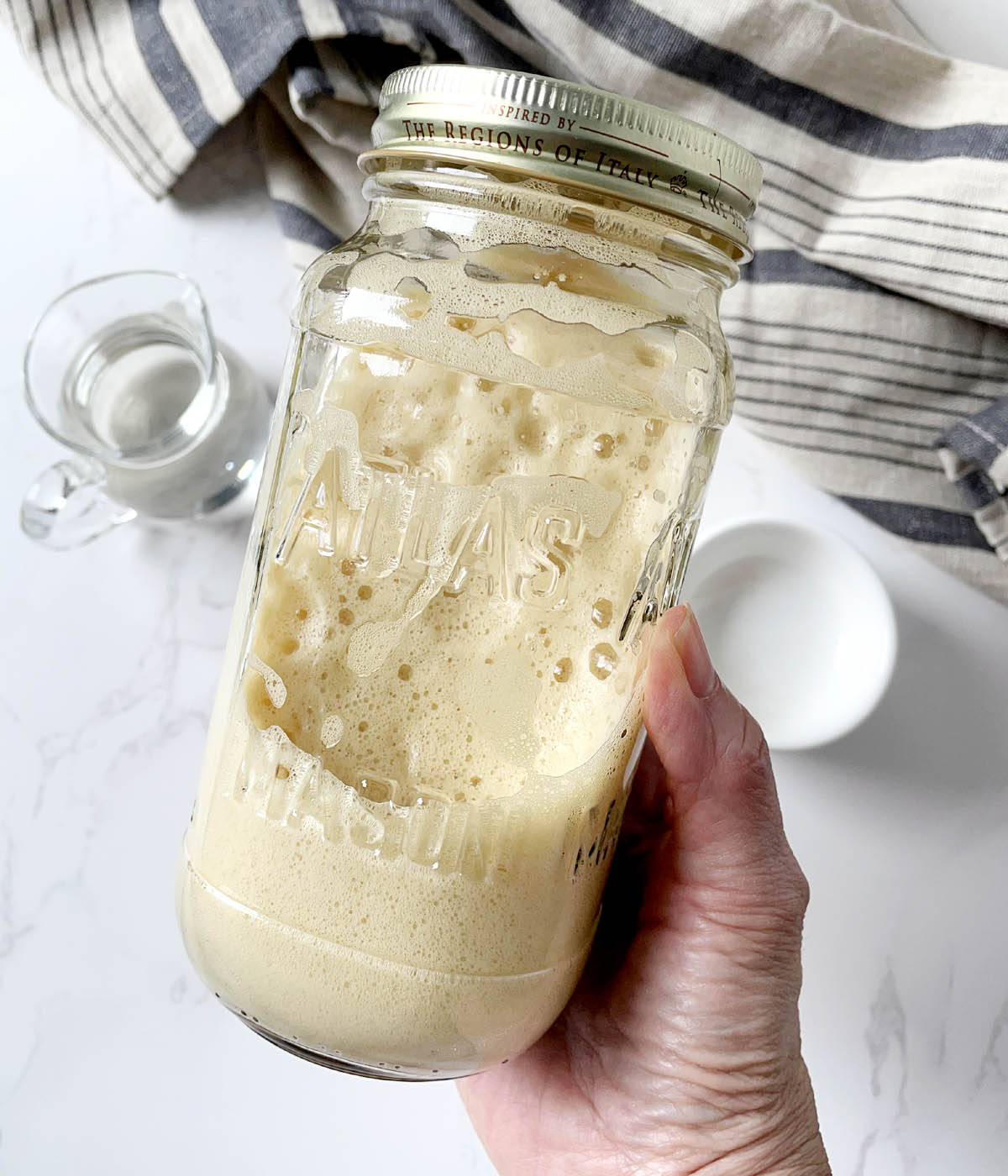 A hand holding a glass jar containing bubbly light brown foam.