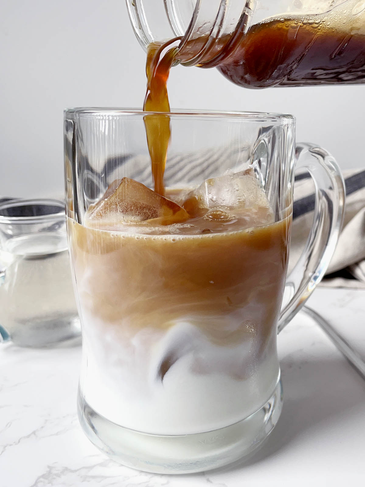 Close-up of a brown liquid being poured into a glass mug containing white milk and ice cubes.