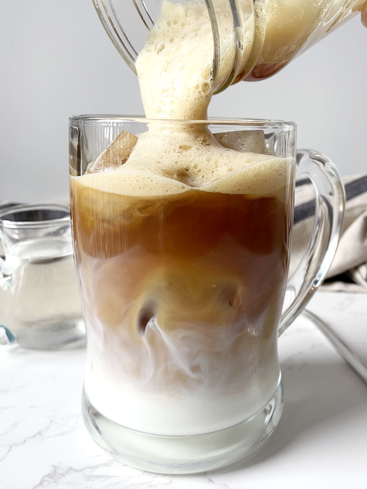 Light brown foam being poured from a glass jar into a glass mug containing white milk and brown coffee.