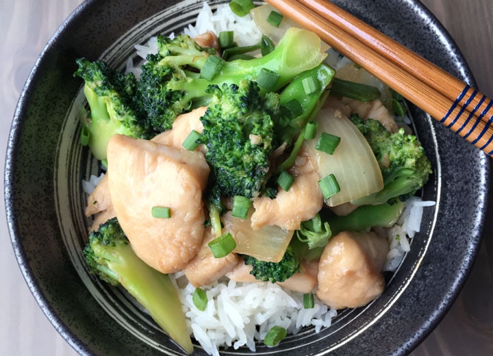 A pair of brown chopsticks on a round bowl containing white rice, green broccoli, and yellow chicken pieces