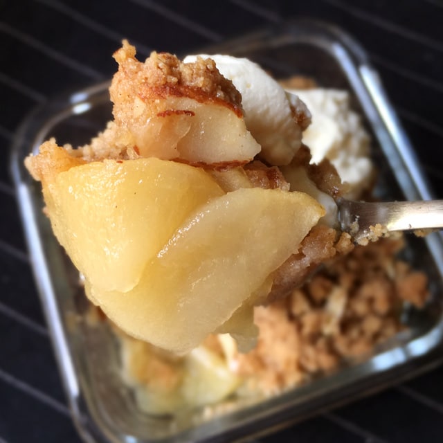 Close-up of a spoonful of yellow cooked apple slices, brown almond crumbs, and white whipped cream