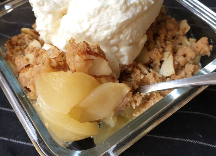 A silver spoon scooping out cooked apples and a crumbly topping with almonds and white whipped cream from a glass square dish