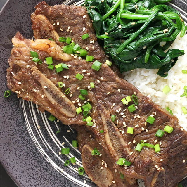A dark round plate containing brown cooked beef short ribs, green vegetables, and white rice, topped with chopped green onion and sesame seeds