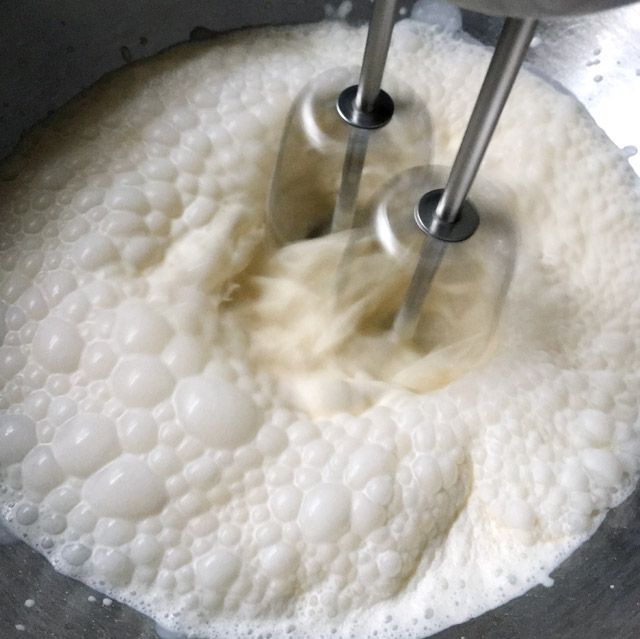 Two metal mixer beaters stirring up bubbles in white cream in a bowl