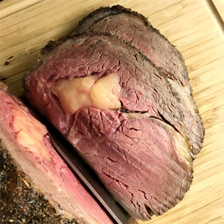 A knife slicing through a beef rib roast, slices of pink and brown beef on a wooden cutting board