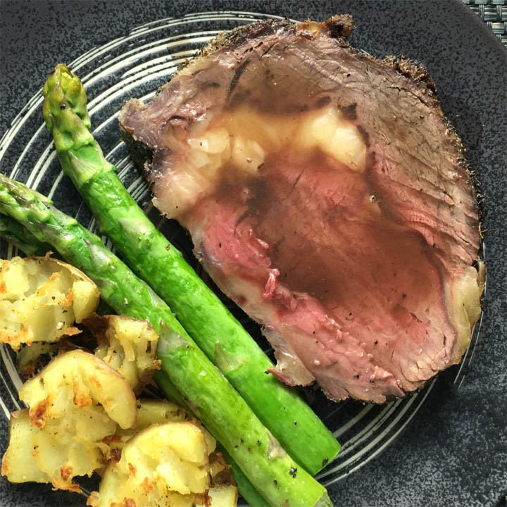 A slice of roast beef, green asparagus spears, and crusty potatoes on a dark round plate