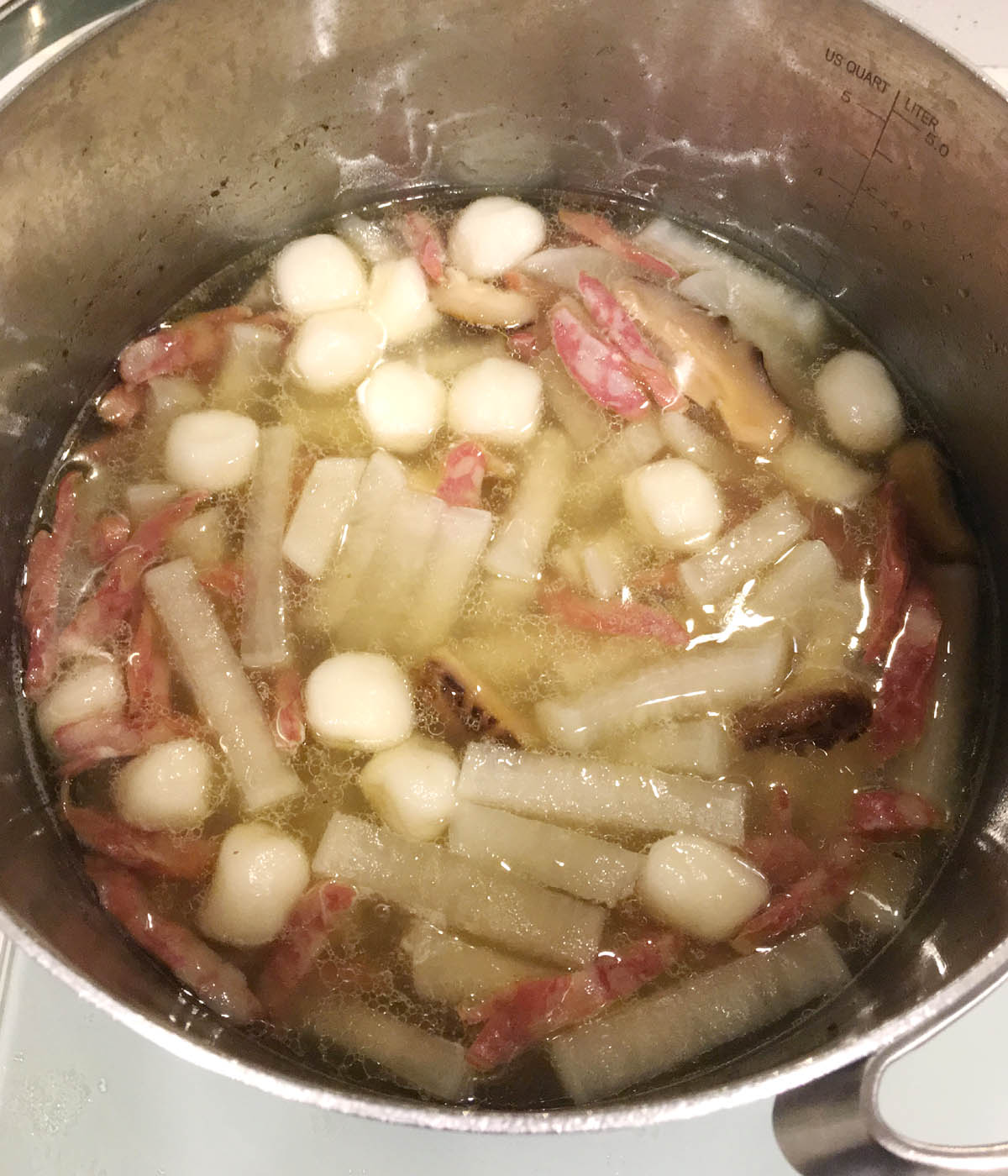 A metal pot containing white dough balls, pink sausage and radish strips in soup.