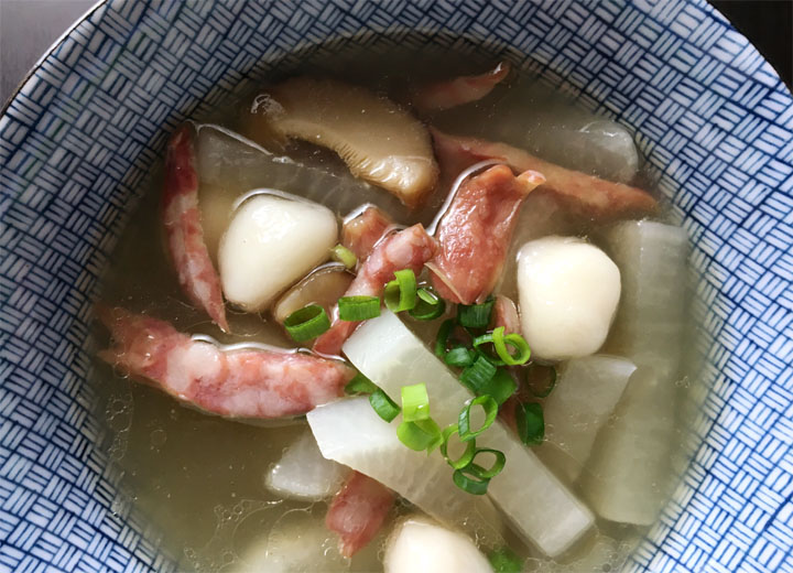 A round blue bowl of Chinese winter solstice soup containing white dough balls, red sausage, chopped green onions, sliced mushrooms, and radish in soup