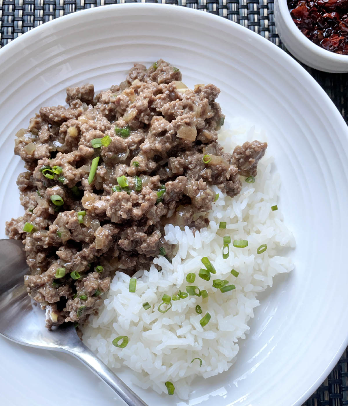 A round white bowl containing a metal spoon, white cooked rice, cooked ground beef, chopped green onions.