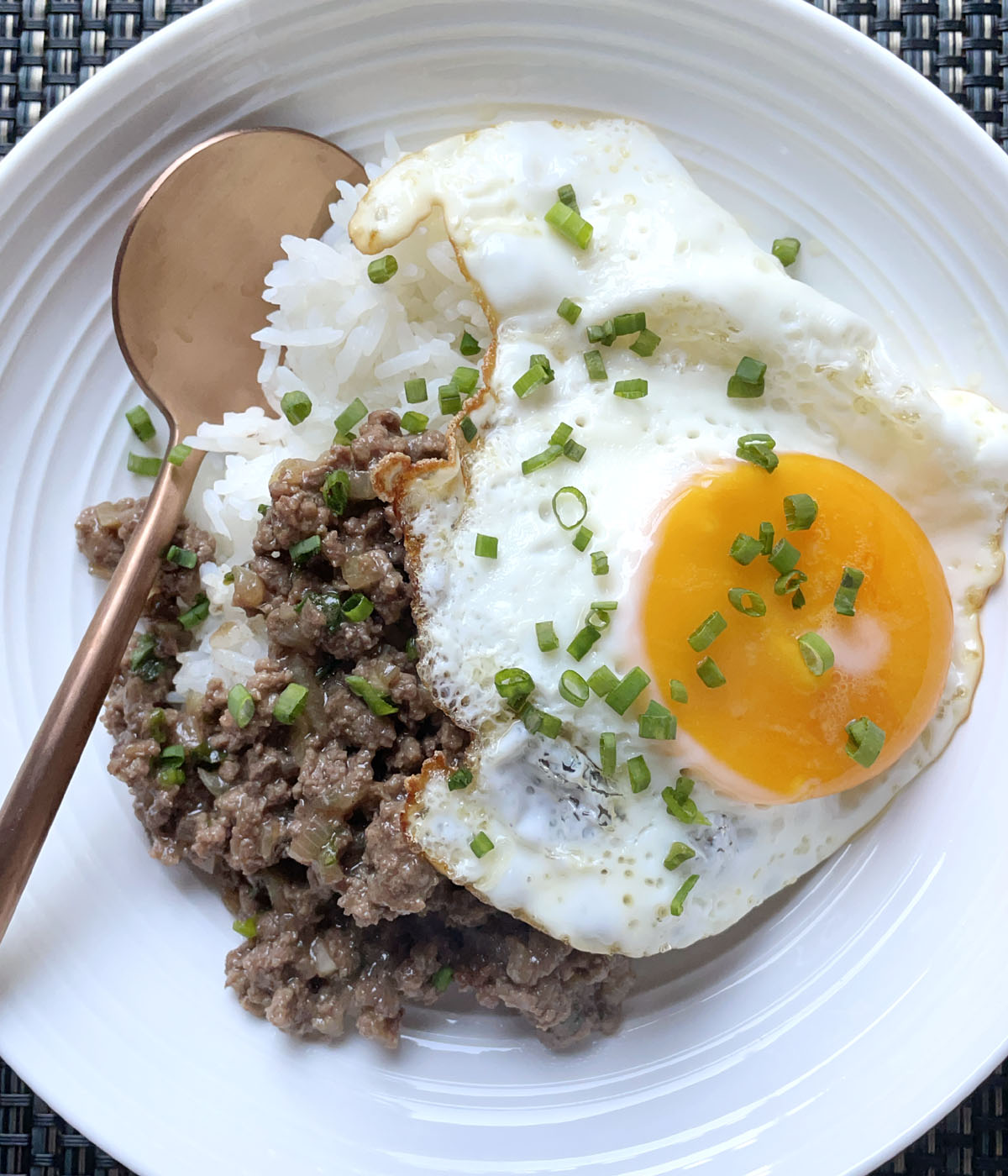 A round white dish containing a bronze colored spoon, white cooked rice, ground meat, and a sunny-side up fried egg, topped with chopped green onions.