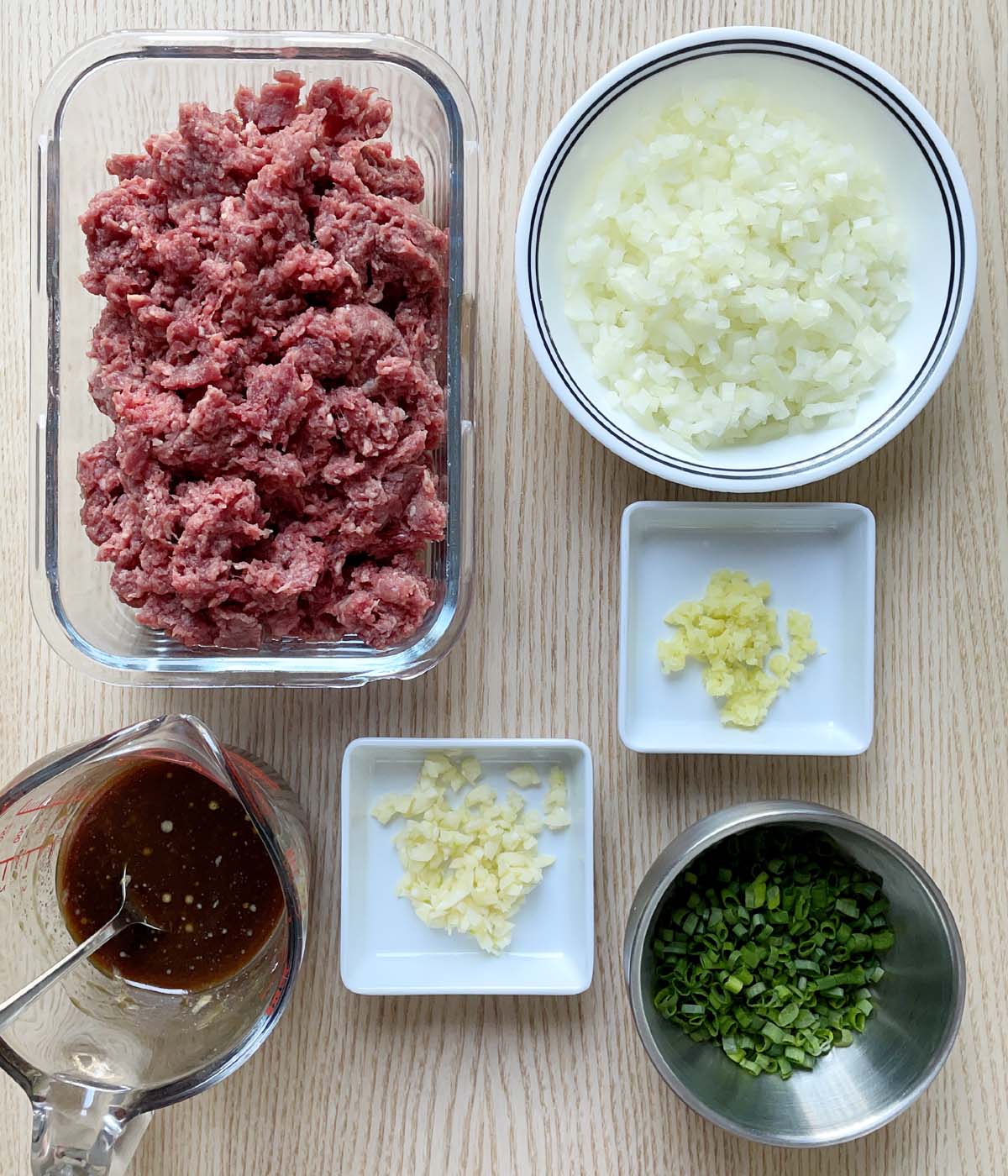 Raw ground meat, a measuring cup with brown sauce, a bowl of diced onions, a dish of minced ginger, a dish or minced garlic, a bowl of chopped green onions.