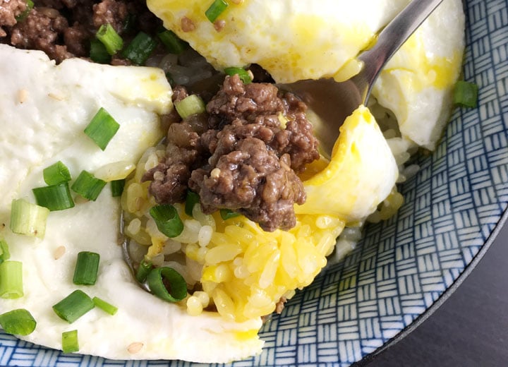 Closeup of a spoon scooping brown ground beef, yellow egg yolk and chopped green onions from a blue and white bowl