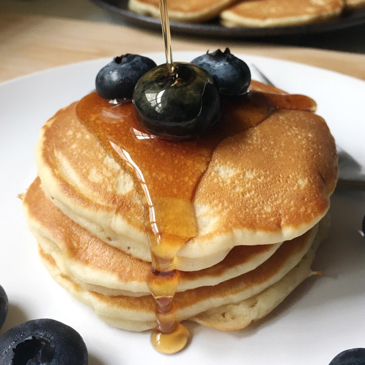 Syrup being poured over a stack of pancakes and blueberies on a white plate