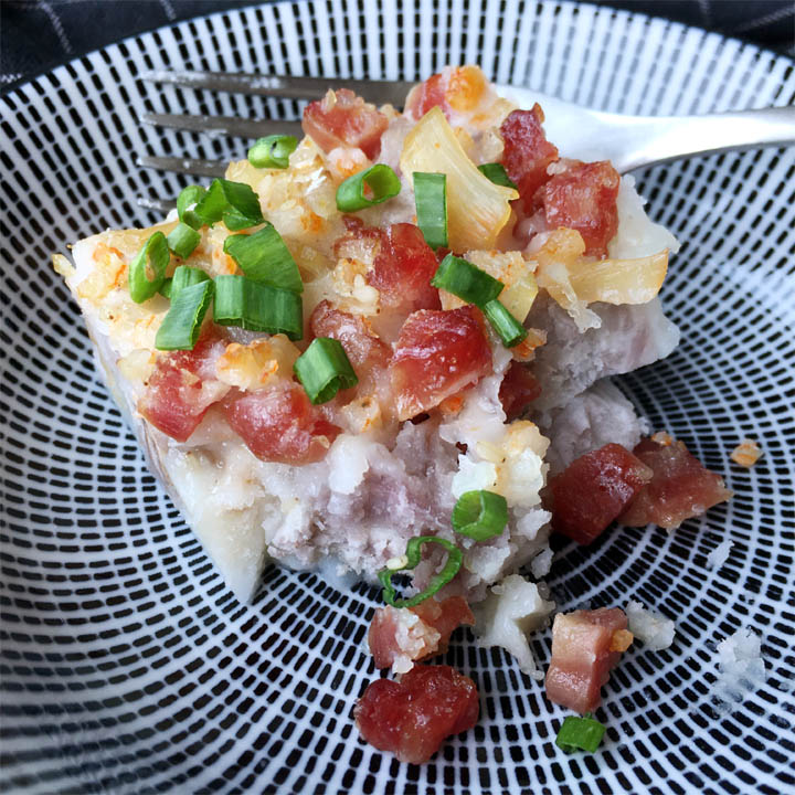 A fork resting behind a piece of taro cake with chopped red sausage, orange seafood, and green onions on a round white and blue plate
