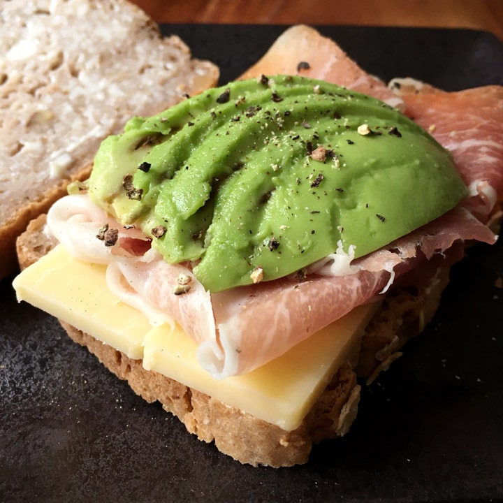 Slices of green avocado, pink ham, and white cheese on a slice of bread on a black plate