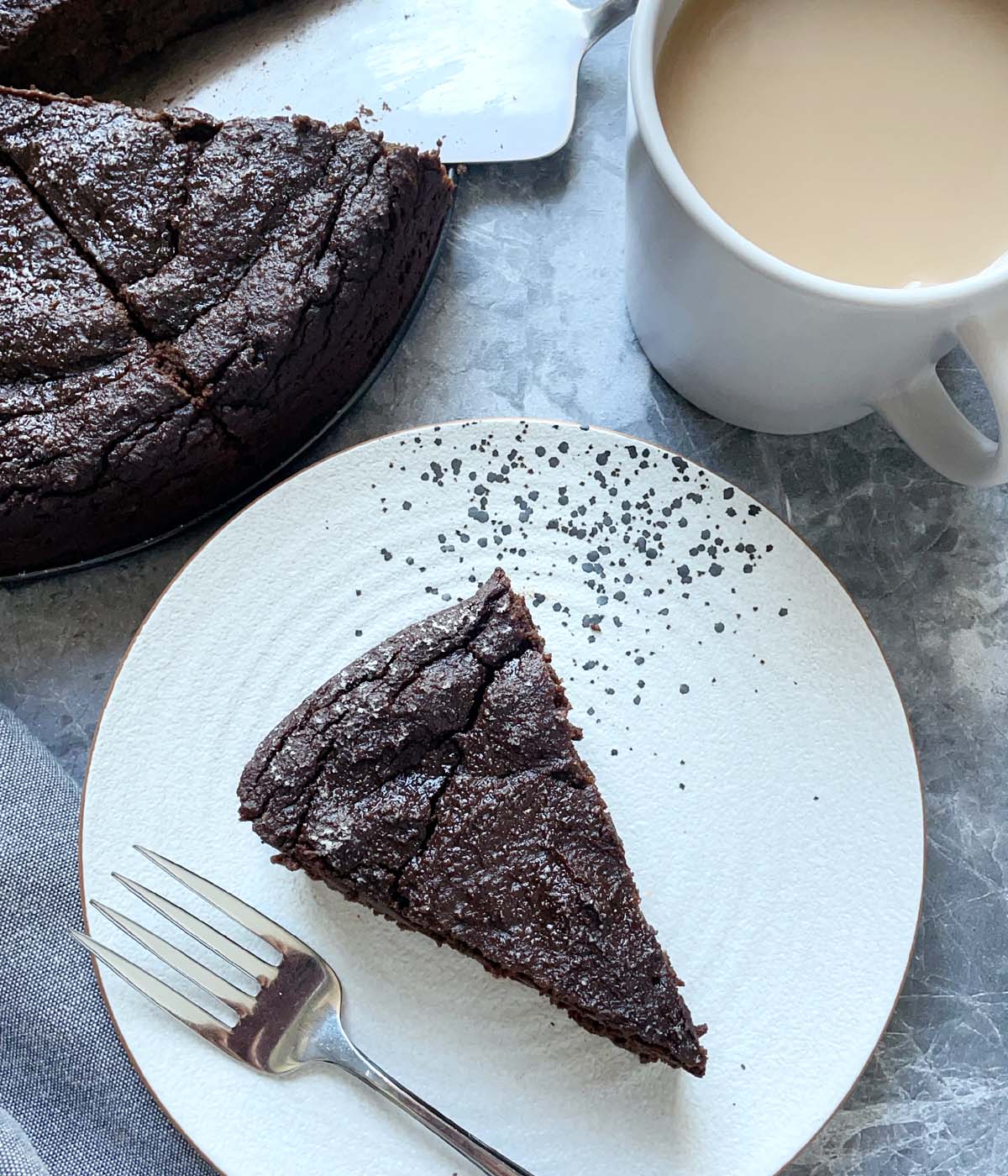 A wedge of dark brown cake on a white plate with a fork next to a cup of tea and chocolate cake.